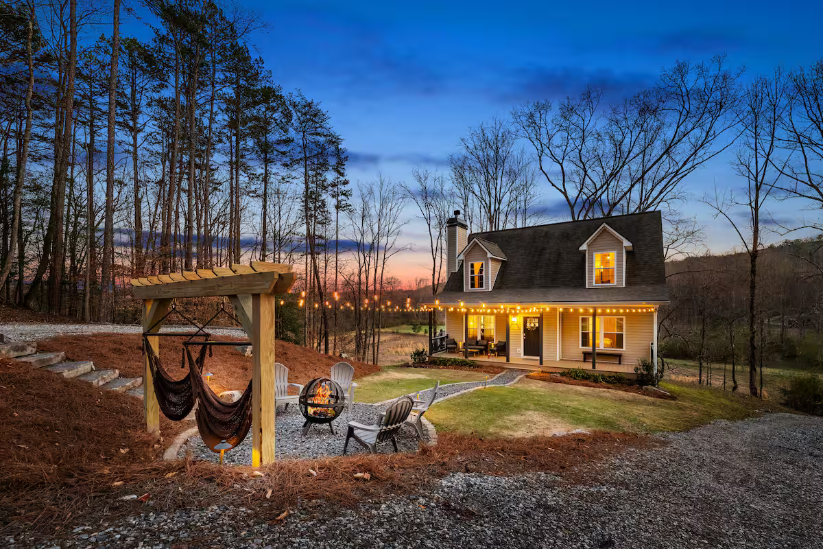 A cozy house with lights strung along the porch, surrounded by trees, with a backyard fire pit area and a hammock on a wooden stand at dusk.