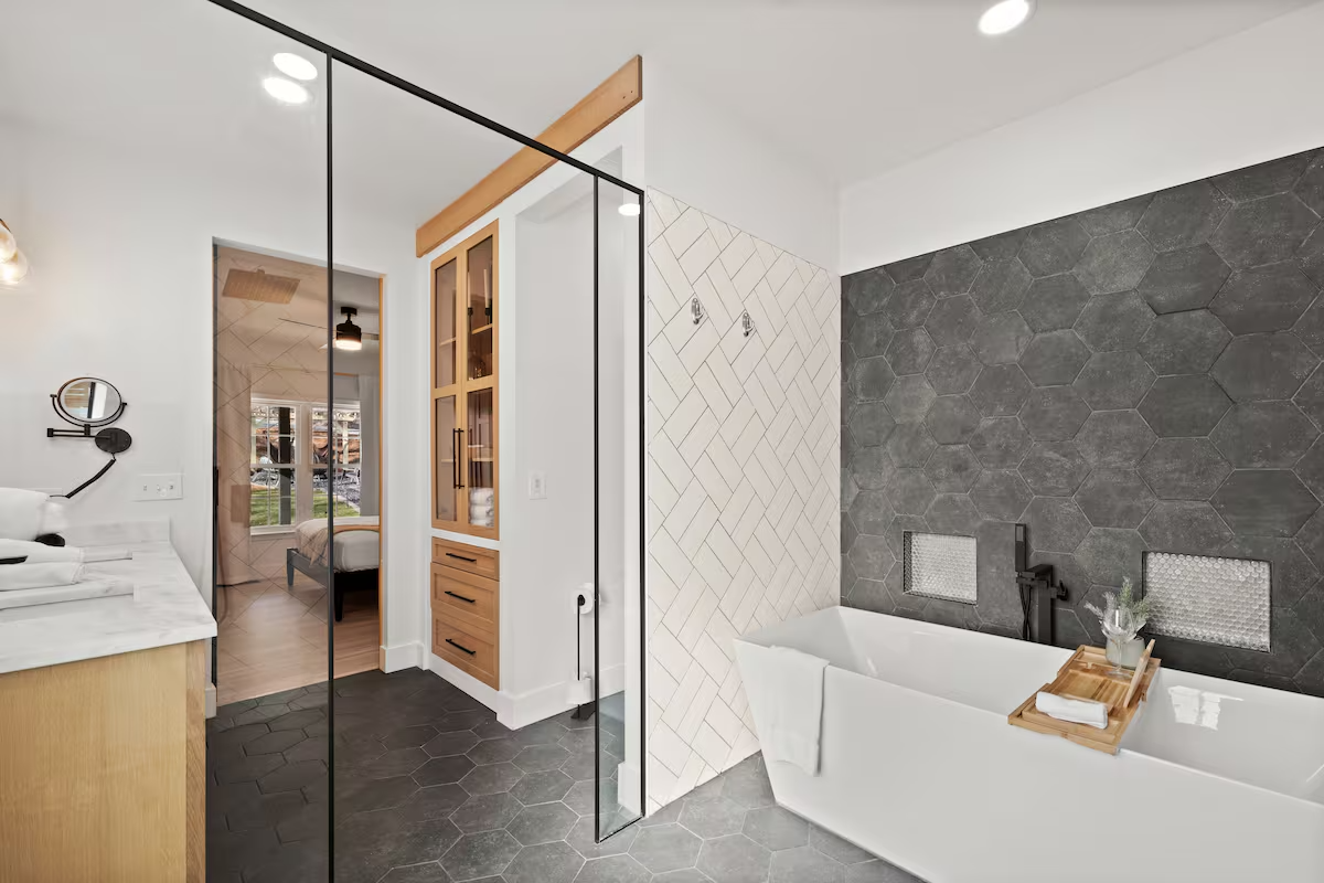 Modern bathroom with dark gray hexagonal tiled walls, a white freestanding bathtub with a wooden tray, and a glass shower enclosure with white herringbone tiles.