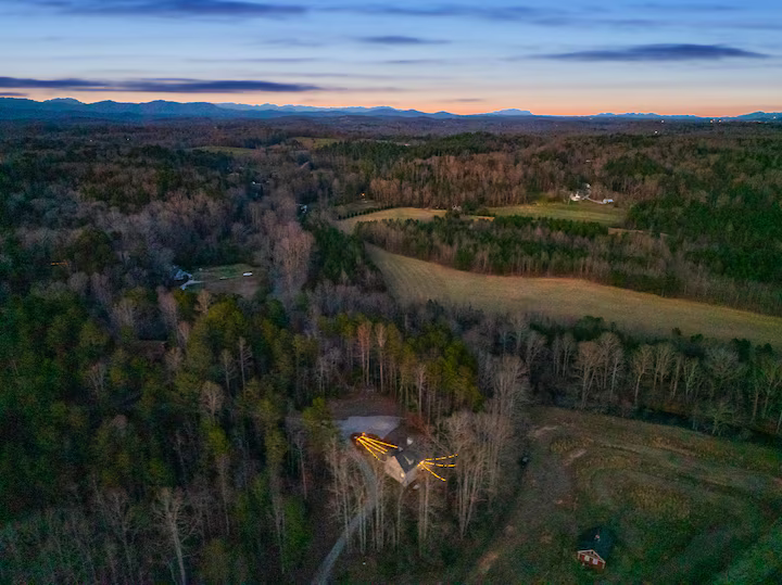 Aerial view of a rural landscape at dusk, showing a dense forest, open fields, distant mountains, and a house with illuminated outdoor lighting.