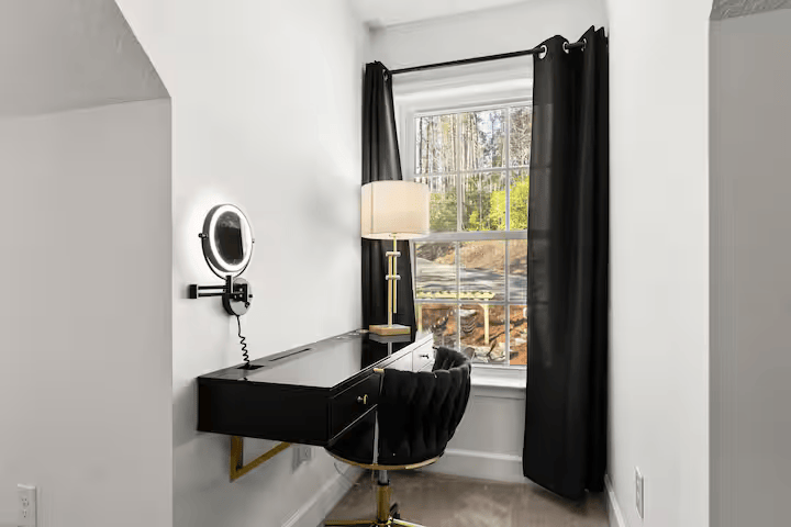 A home office corner featuring a black desk, black swivel chair, a small table lamp, a wall-mounted mirror with a light, and a window with black curtains.
