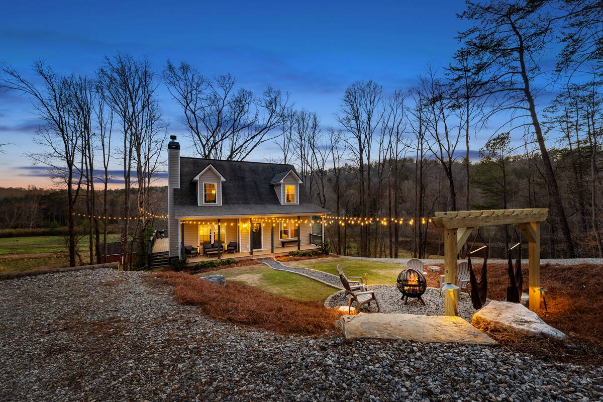 A cozy home with a front porch decorated with string lights, surrounded by trees, at dusk. There is a gravel driveway, a small yard with chairs, a fire pit, and a wooden pergola with hanging fabric and a lantern.