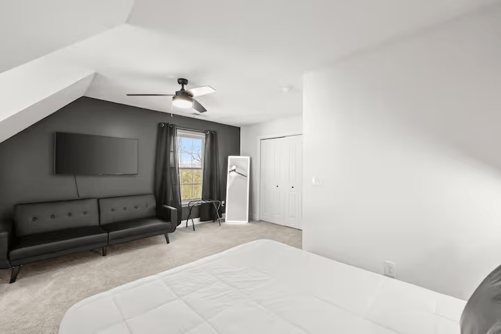Modern bedroom with white walls, a black sofa, a wall-mounted TV, a ceiling fan, a window with gray curtains, a full-length mirror, and a door.