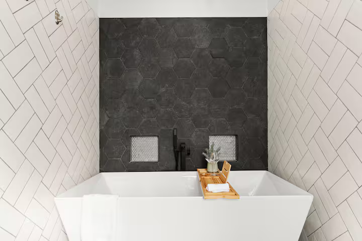Bathtub with a black faucet, a wooden tray with a plant and towel, against a wall with dark hexagonal tiles and white rectangular tiles in a bathroom.