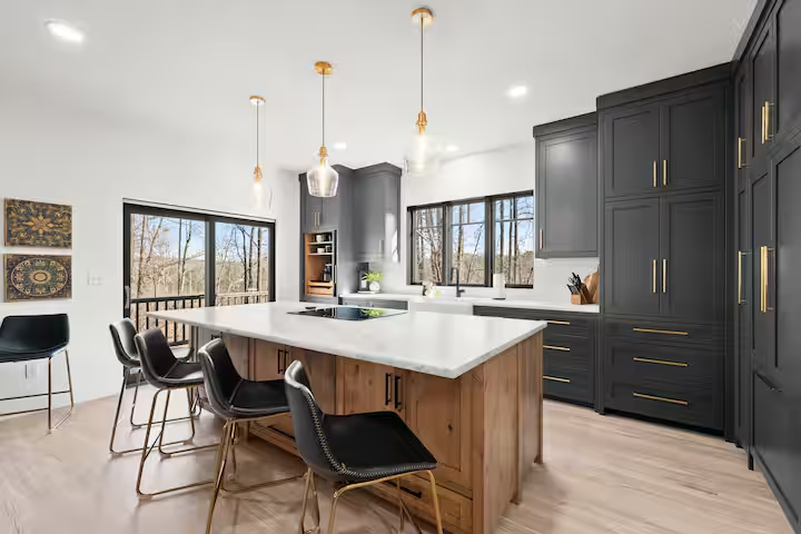 Modern kitchen with dark cabinets, a white island with seating, pendant lights, and large windows showing an outdoor view.