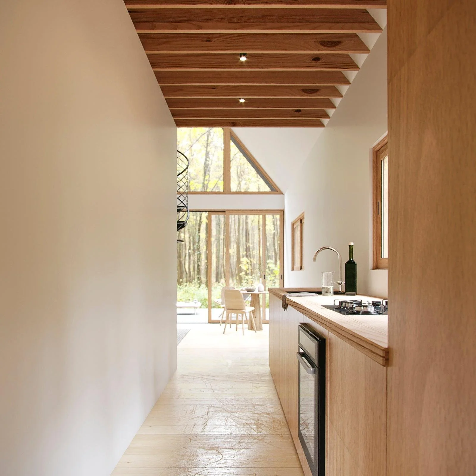 A modern kitchen with wooden cabinets, a countertop with a sink, and a built-in oven. Large windows and a glass door lead to an outdoor area with trees, allowing natural light to fill the space.