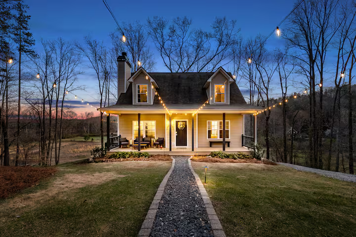 A cozy house on a hill during dusk with string lights hanging above, trees in the background, and a stone pathway leading to the front door.