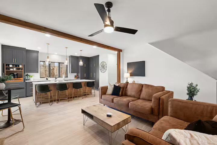 Open-concept living room and kitchen with modern decor, brown couches, a coffee table, ceiling fan, and large window.