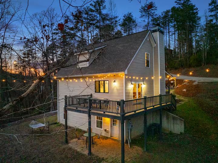 A modern two-story house with beige siding, a deck with black railing, string lights, and large windows, surrounded by trees at dusk.