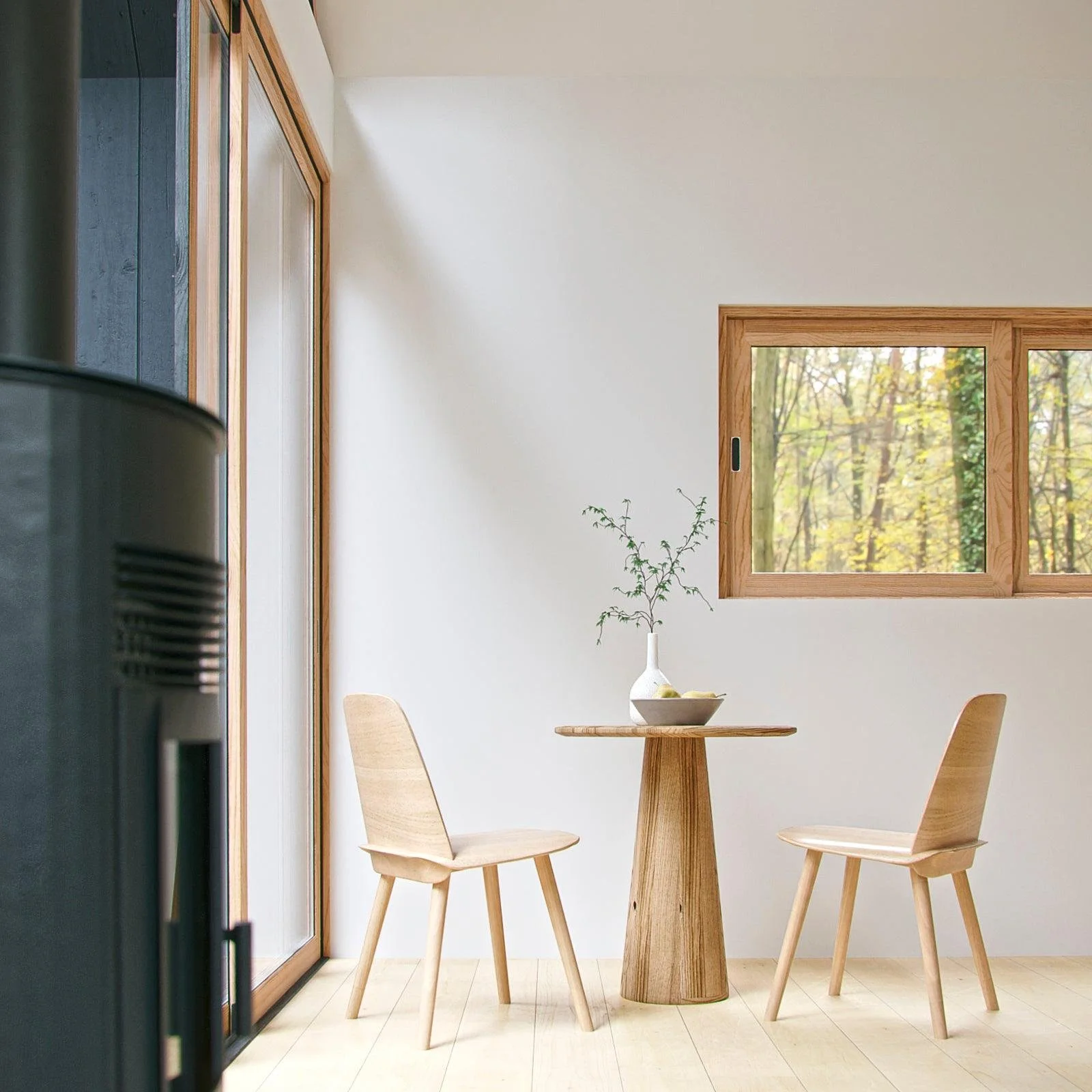 Minimalist dining nook with a small round wooden table, two wooden chairs, a white vase with branches, and a window showing trees outside in a bright room.