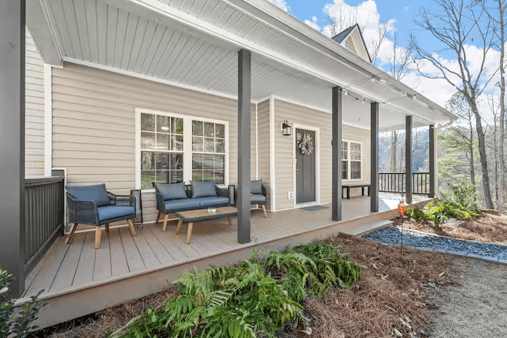Covered front porch of a house with outdoor seating, beige siding, black support beams, and view of trees.