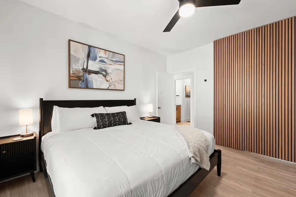 A modern bedroom with a large bed, white bedding, black headboard, small black nightstands with lamps, abstract artwork above the bed, a wooden slatted room divider, and hardwood floors.
