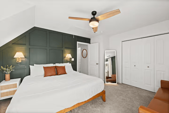 Bedroom with a large bed, dark green accent wall, wall-mounted lamps, ceiling fan, full-length mirror, white closet doors, and a brown armchair.