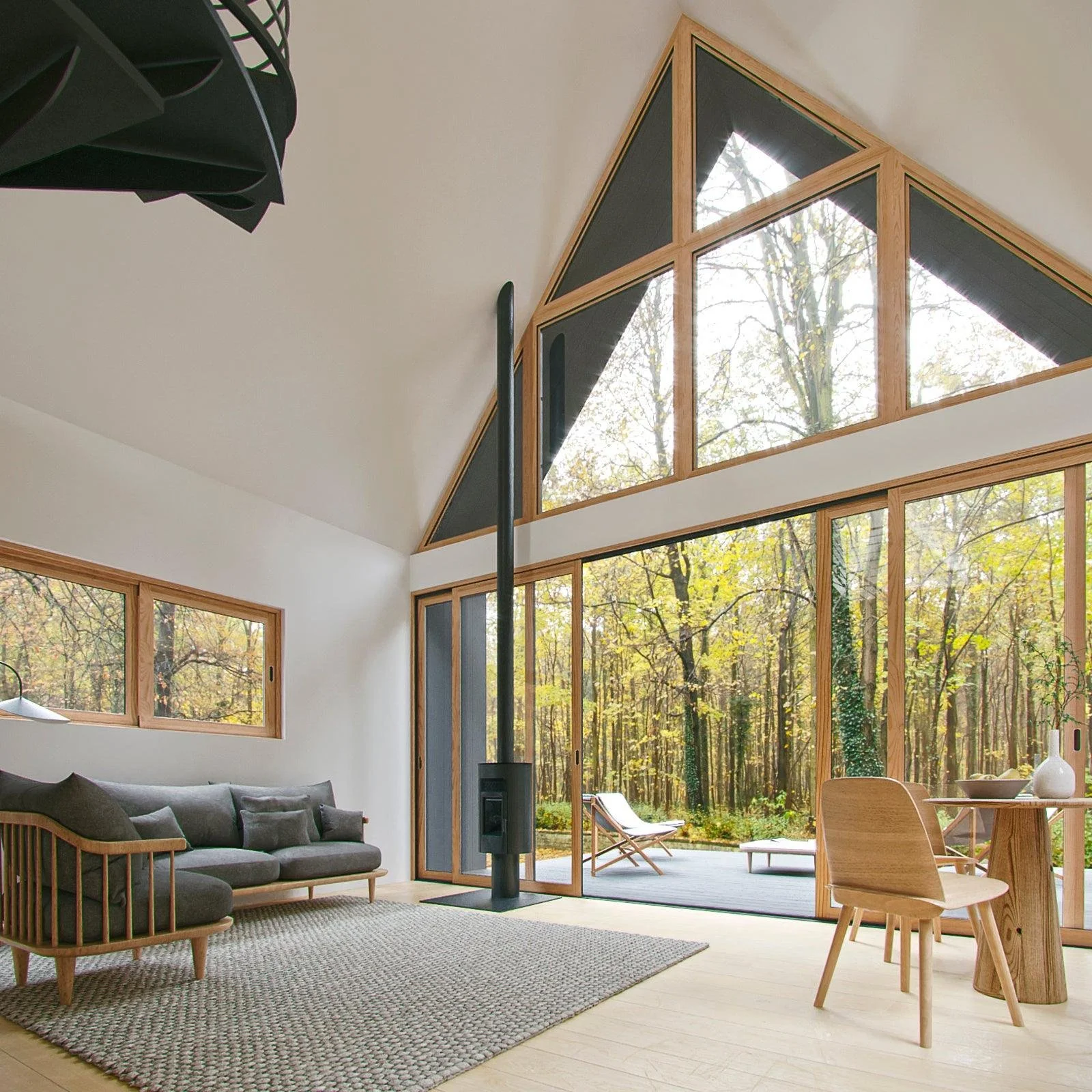 A modern living room with large floor-to-ceiling windows showing a forest outside. The room has a gray sofa, a small wooden table and chair, a wood-burning stove, and a patio with outdoor furniture visible through glass doors.