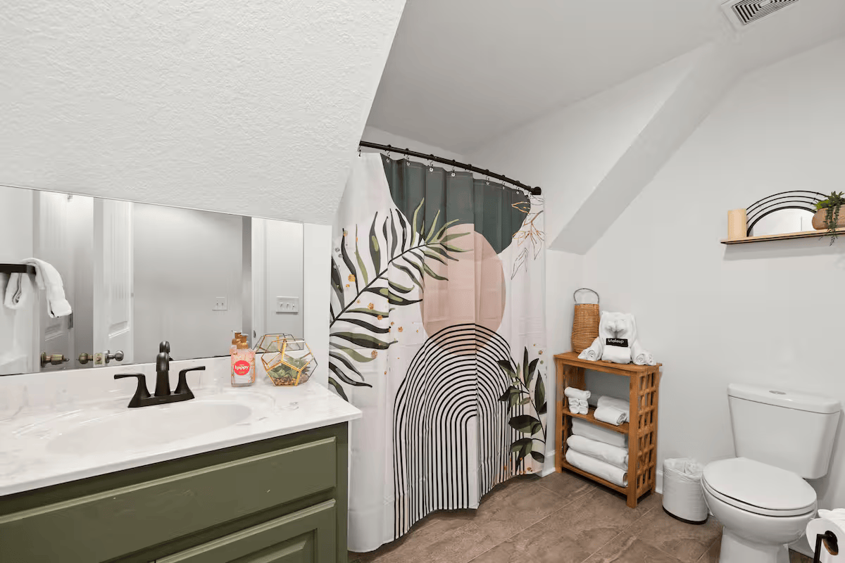Bathroom with a green vanity, a white countertop, a large mirror, a decorative shower curtain with abstract and botanical patterns, a wooden shelf with towels and decor, a toilet, and a small trash bin.