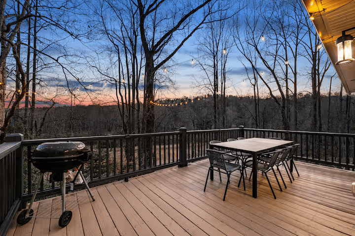 Empty wooden deck with a table and chairs, a barbecue grill, string lights, and a view of a wooded landscape at sunset.