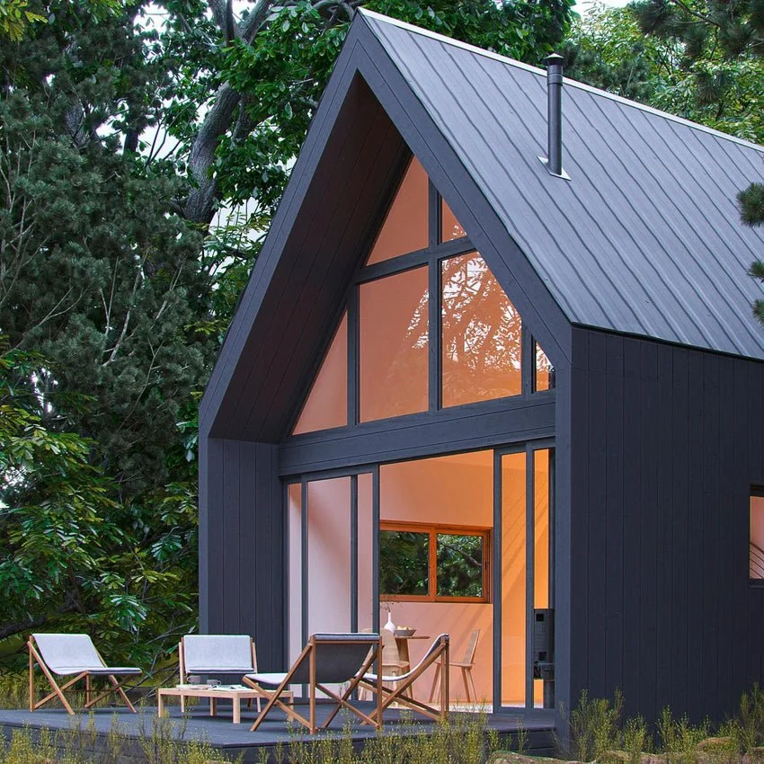 A modern, black, A-frame house with large glass windows surrounded by green trees, with an outdoor seating area featuring chairs and a table.