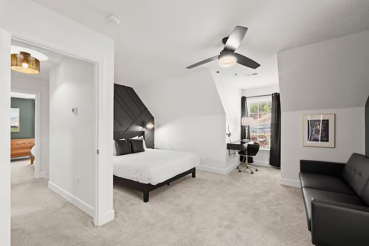 Modern bedroom with white walls, black accent wall behind bed, white bedding, black furniture, ceiling fan, window with curtains, small workspace with desk and chair, black sofa, framed artwork on wall.
