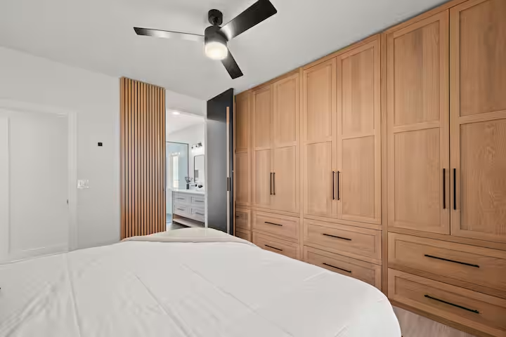 Modern bedroom with a large wooden wardrobe, a ceiling fan, and a view into a bathroom with white cabinetry and a window.