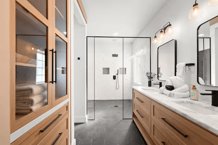 Modern bathroom with wooden vanity, large mirror, black accents, glass shower enclosure, and white tiled walls.