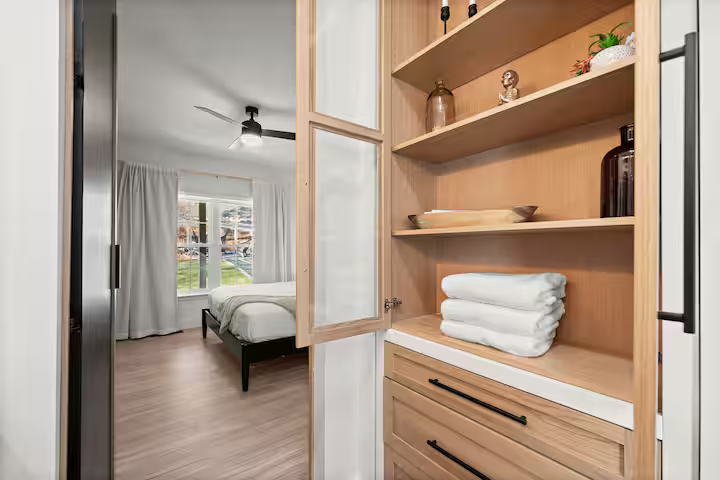 Open pantry with folded towels and decorative items in a bedroom setting with a bed and window in the background.