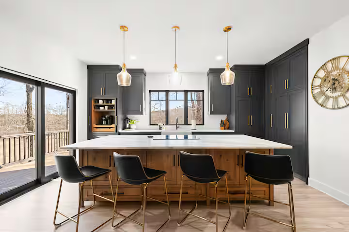 Modern kitchen with black cabinetry, white countertops, and a central island with four black barstools. Pendant lights hang above the island, and large windows and sliding glass door provide natural light, with a view of a deck and trees outside.