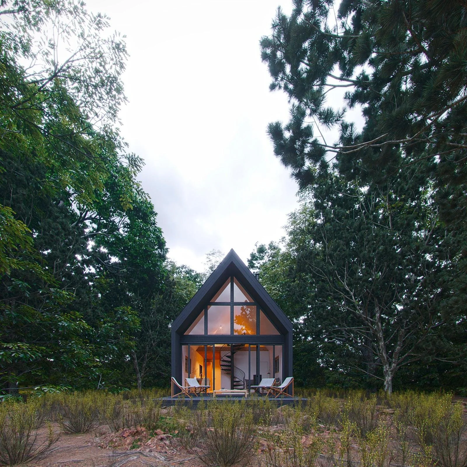 A modern A-frame cabin with large glass windows, surrounded by trees, with an outdoor seating area on a grassy clearing.