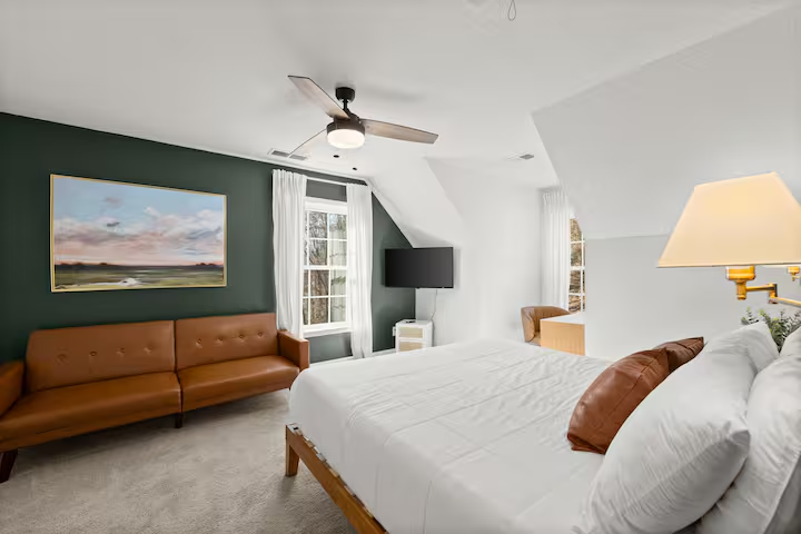 Clean, modern bedroom with white walls, brown leather sofa, bed with white linens and brown pillows, wall-mounted TV, picture of a landscape, window with white curtains, and a ceiling fan.