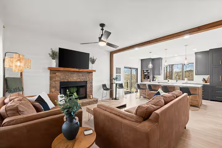 Open-concept living room and kitchen with brown leather sofas, a brick fireplace with a mounted TV, a ceiling fan, modern pendant lights, and large windows showing an outdoor view.