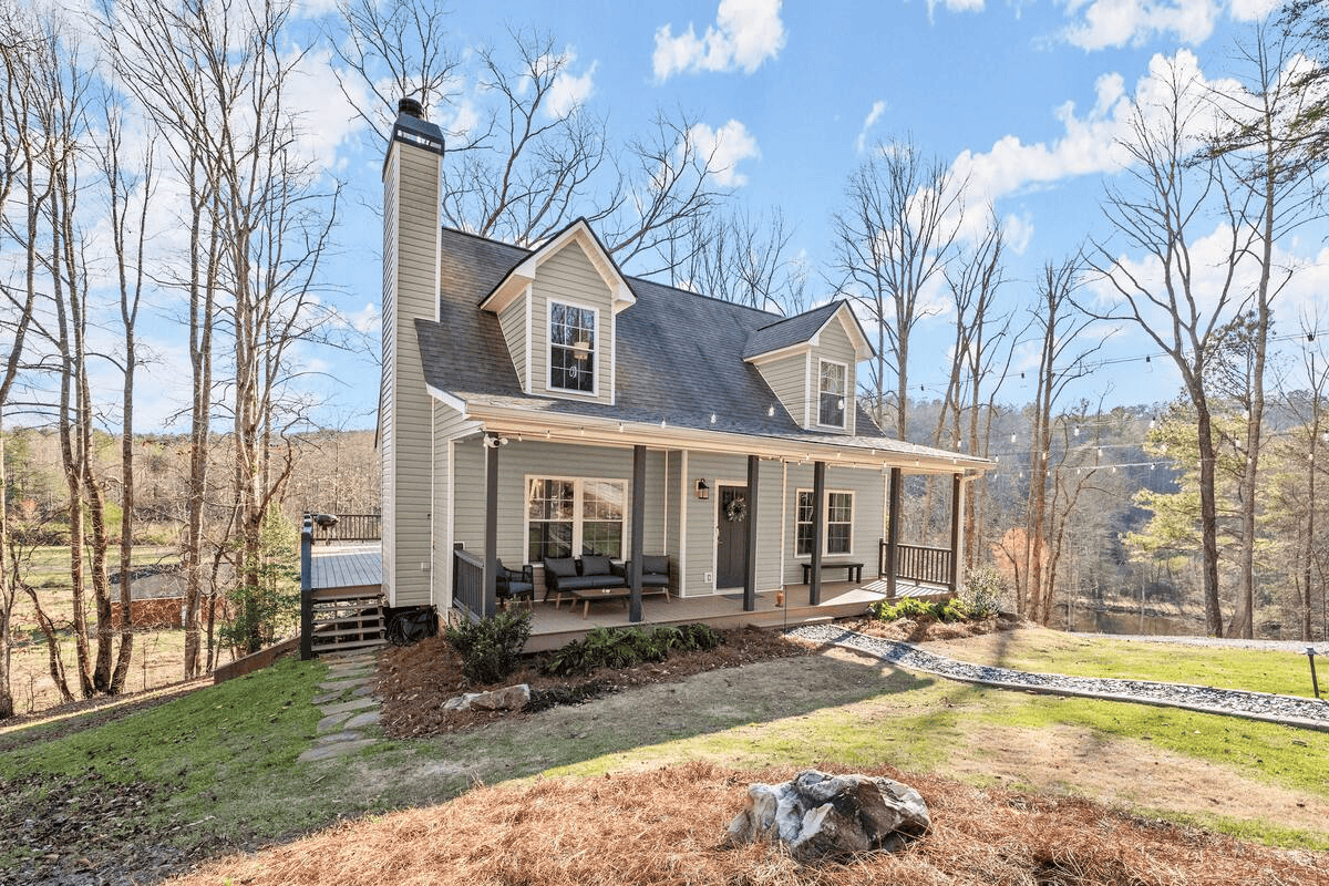 A two-story house with a front porch, a black front door with wreath, and outdoor seating, surrounded by a grassy yard and trees, with a blue sky and white clouds above.