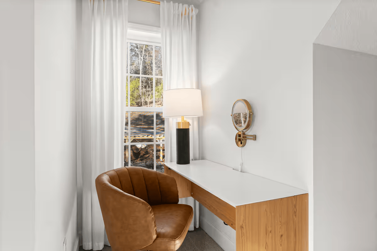 Corner of a room with a small wooden desk, brown leather chair, a table lamp, white curtains, and a window with a view outside.