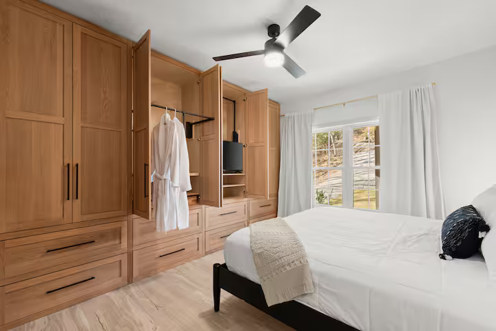 Bedroom with large built-in wooden closet, white bed with a beige blanket, black pillow, ceiling fan, window with white curtains, and light-colored flooring.