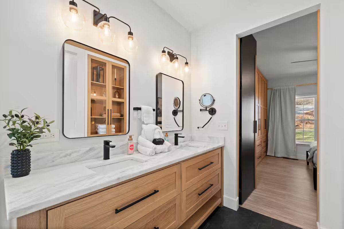 Modern bathroom with double vanity, white marble countertop, wooden cabinets, black fixtures, wall-mounted mirrors, wall-mounted lamps, a mirror with built-in storage, a small potted plant on the left, soaps, and towels, with an open door leading to