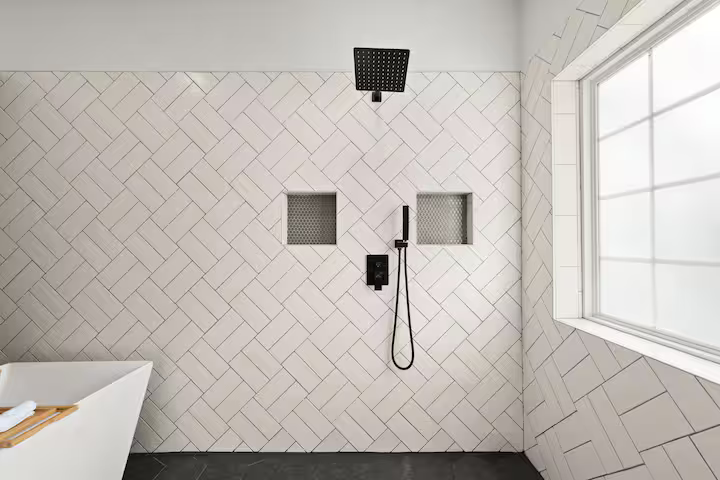 Modern shower area in a bathroom with a black rainfall showerhead, two small textured wall niches, a handheld shower, and large window with frosted glass.