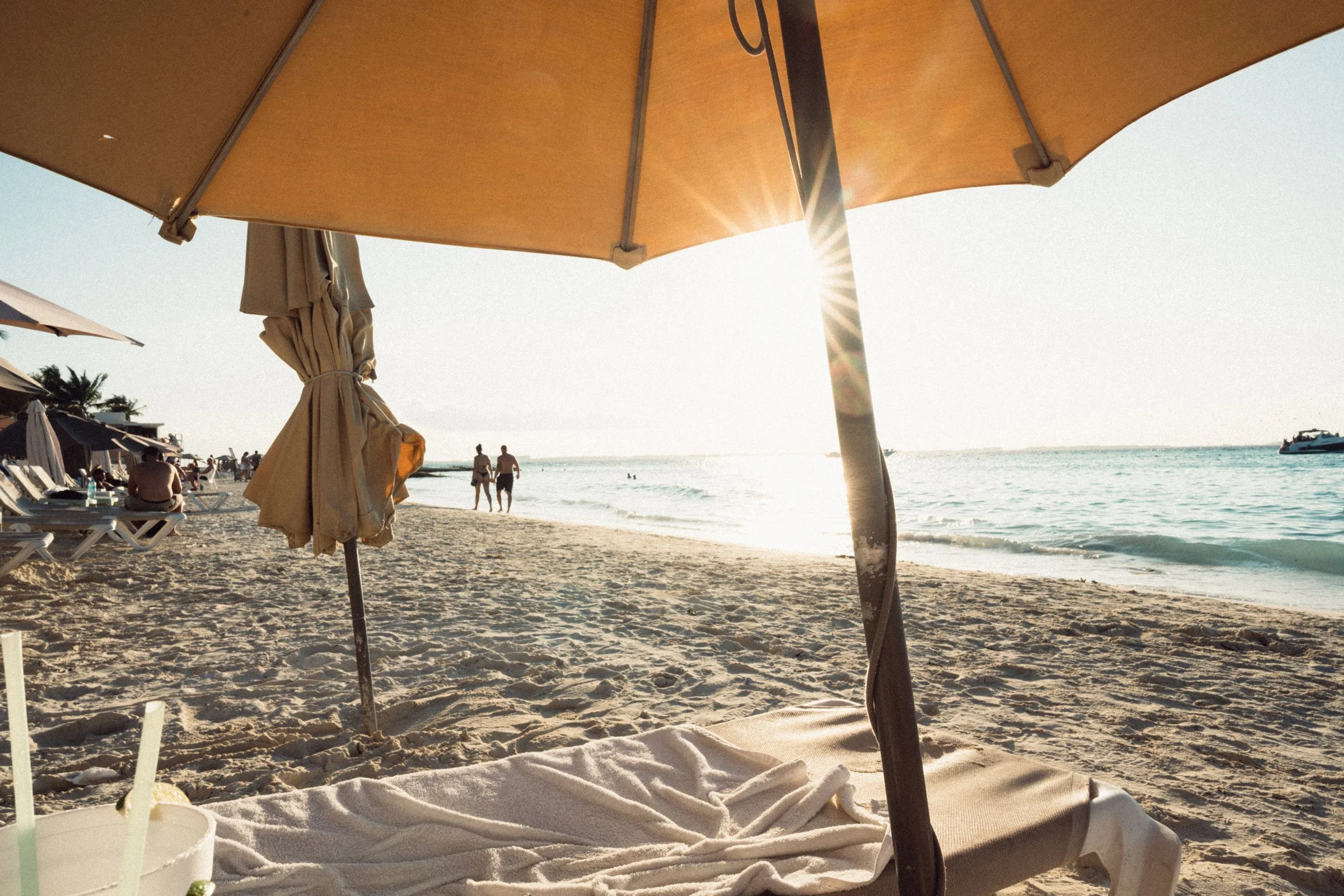 Beach scene with a large beige umbrella, a towel on a lounge chair, and the sun setting over the ocean, with people walking along the shoreline and the ocean with boats in the background.