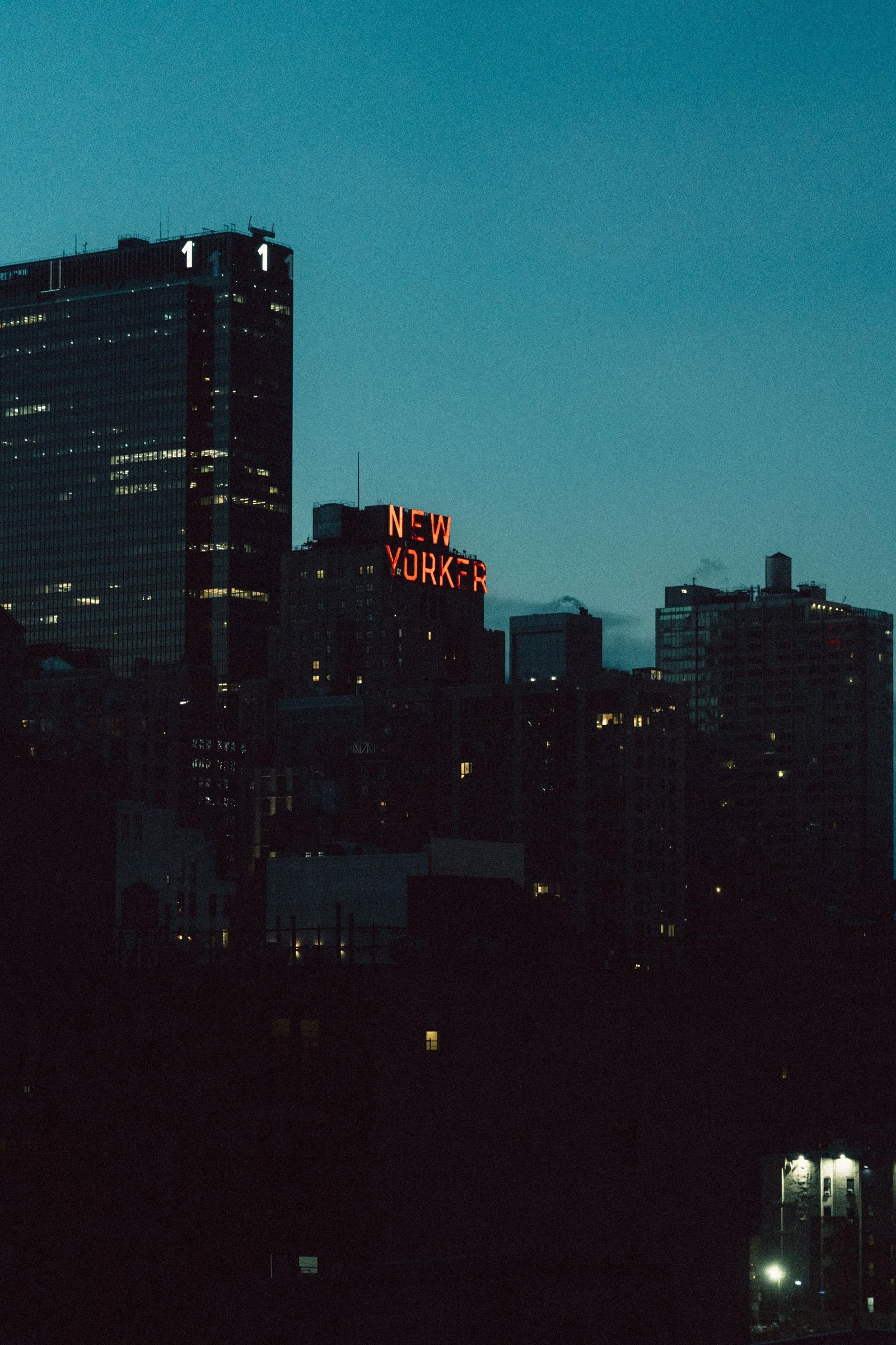 Nighttime cityscape of New York City with illuminated buildings, including a neon sign reading 'NEW YORKER' on one of the skyscrapers.
