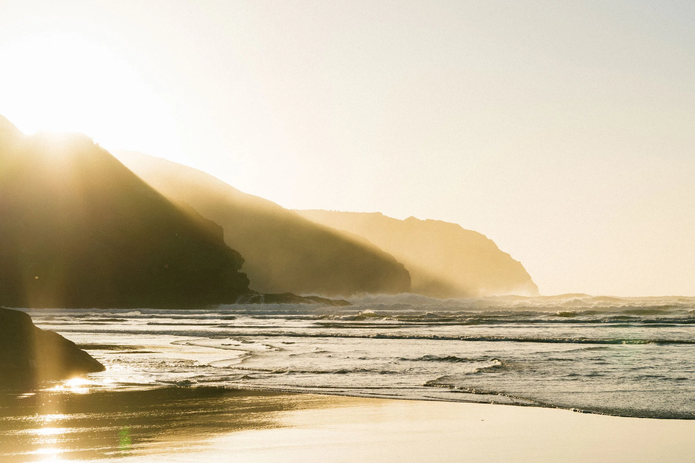 Sunset beach scene with waves crashing on the shore, cliffs in the distance, and sunlight casting a warm glow over the water.