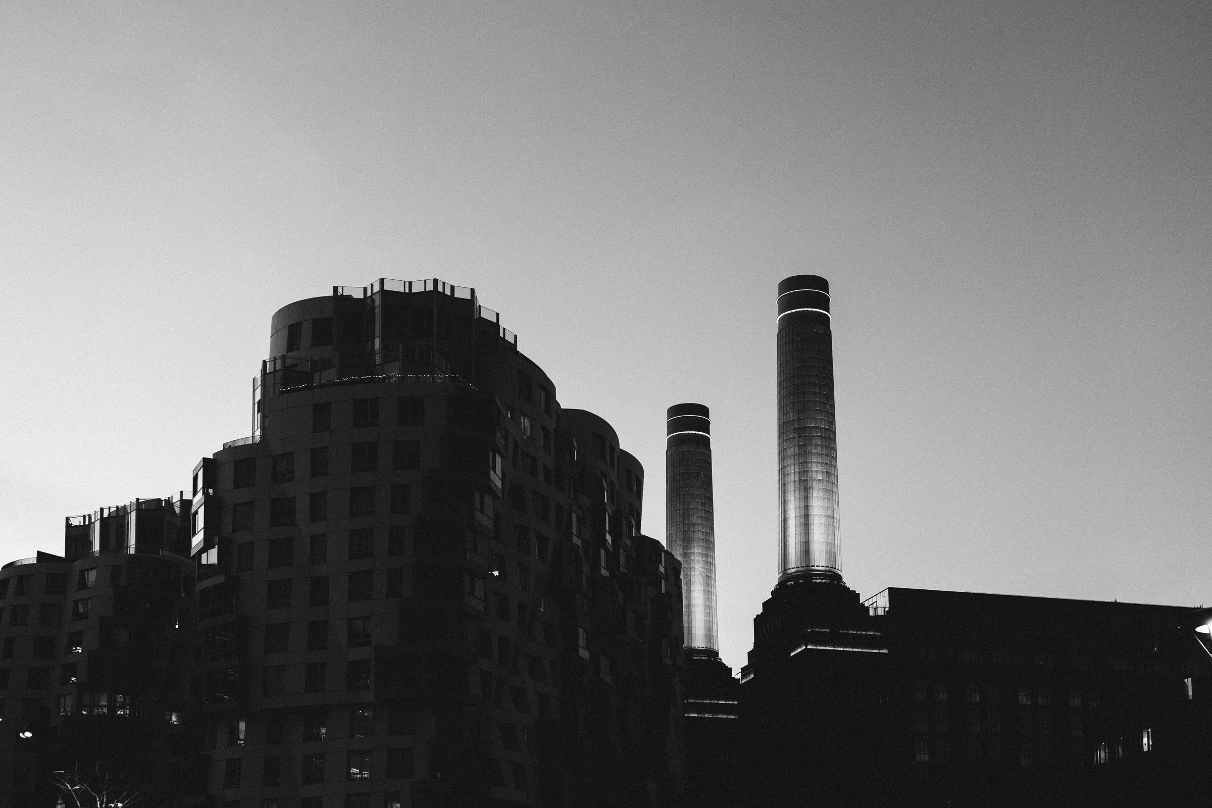 Black and white photo of modern residential buildings with two tall smokestack chimneys in the background, against a clear sky.