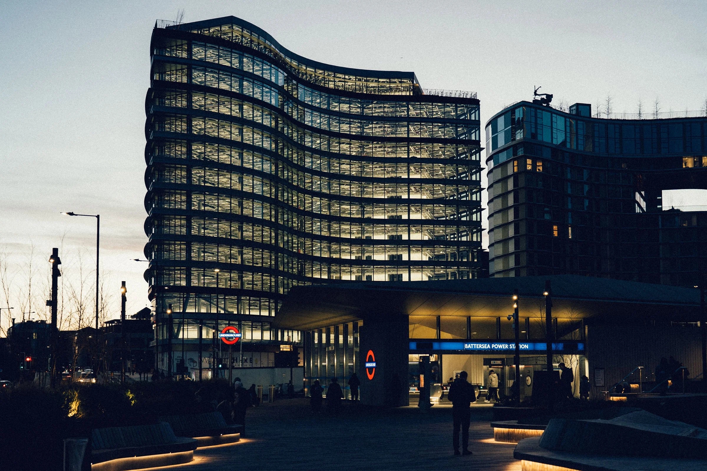 Modern multi-story building with wavy glass exterior, illuminated lobby entrance labeled Battersea Power Station, near London Underground sign, evening setting