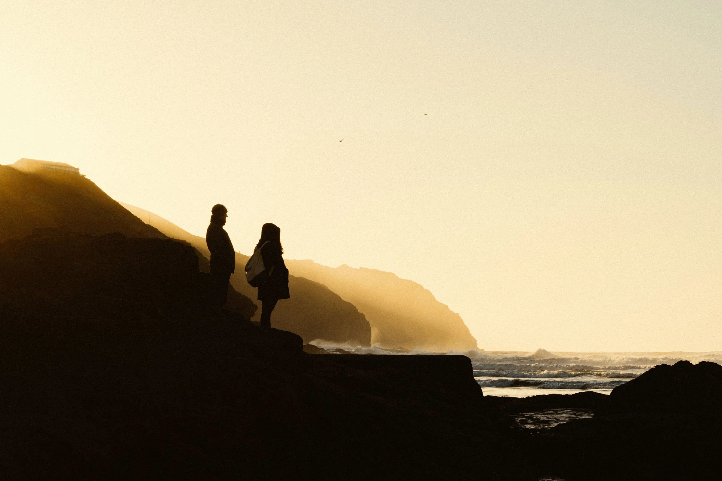 Silhouette of a man and woman standing on a rocky coastline at sunset with cliffs and ocean waves in the background.