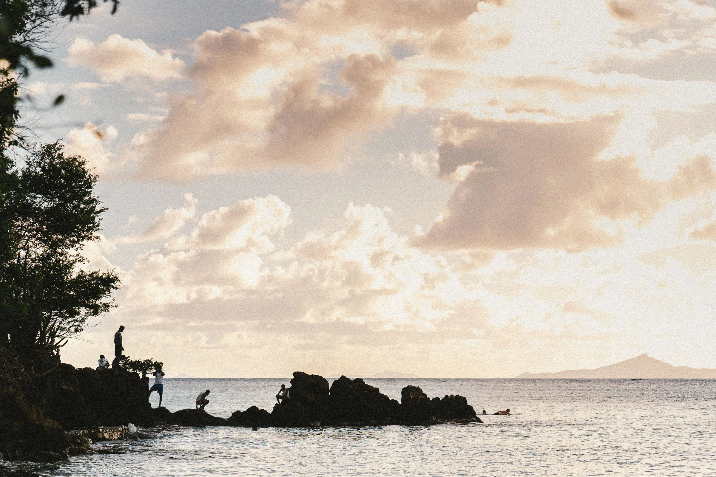 People standing and sitting on rocks by the shoreline under a cloudy sky during sunset or sunrise, with the ocean and distant island in the background.