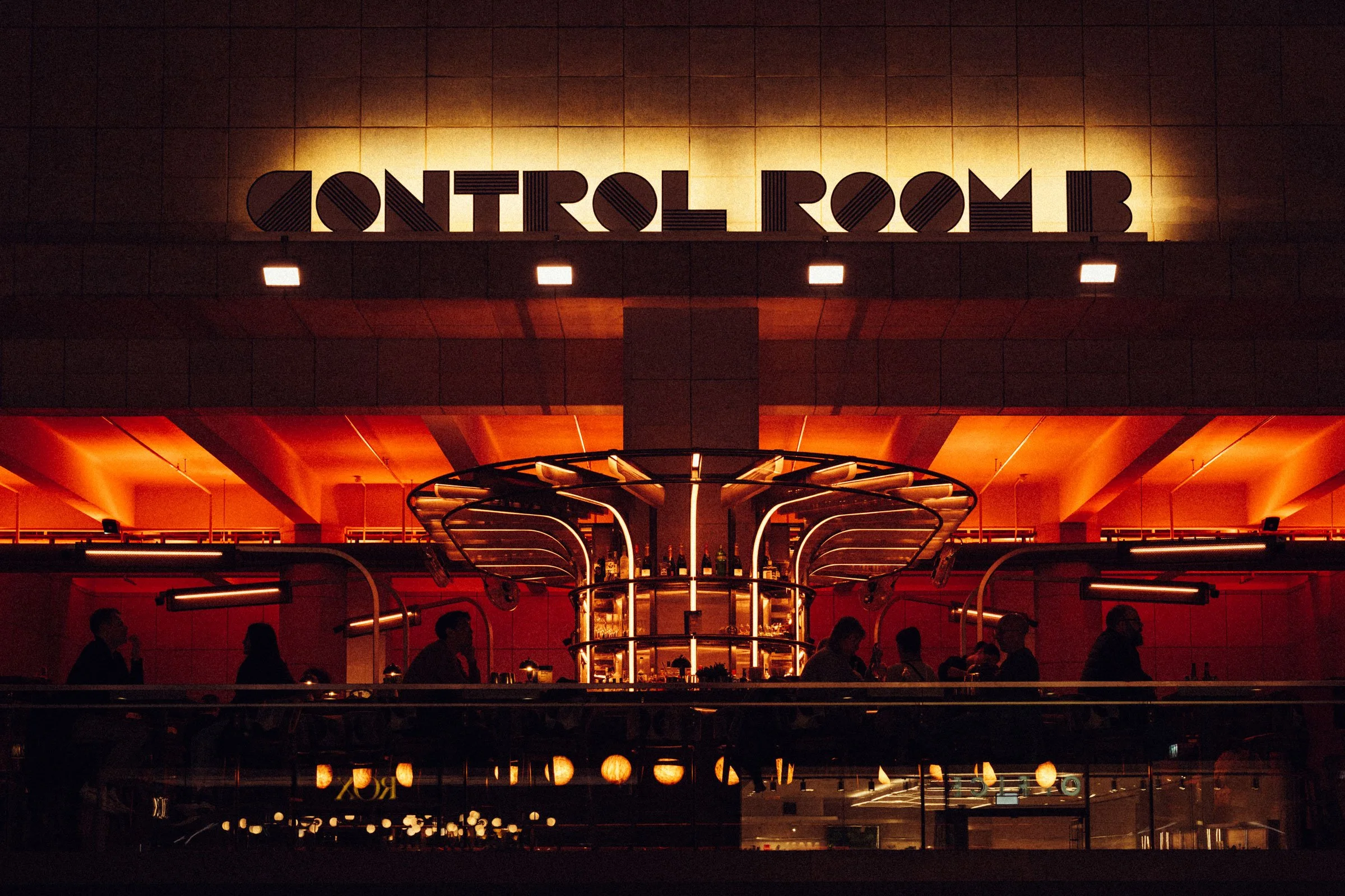 Night view of a bar inside a control room with warm lighting, silhouettes of people sitting and talking, and a modern bar structure with bottles on shelves.