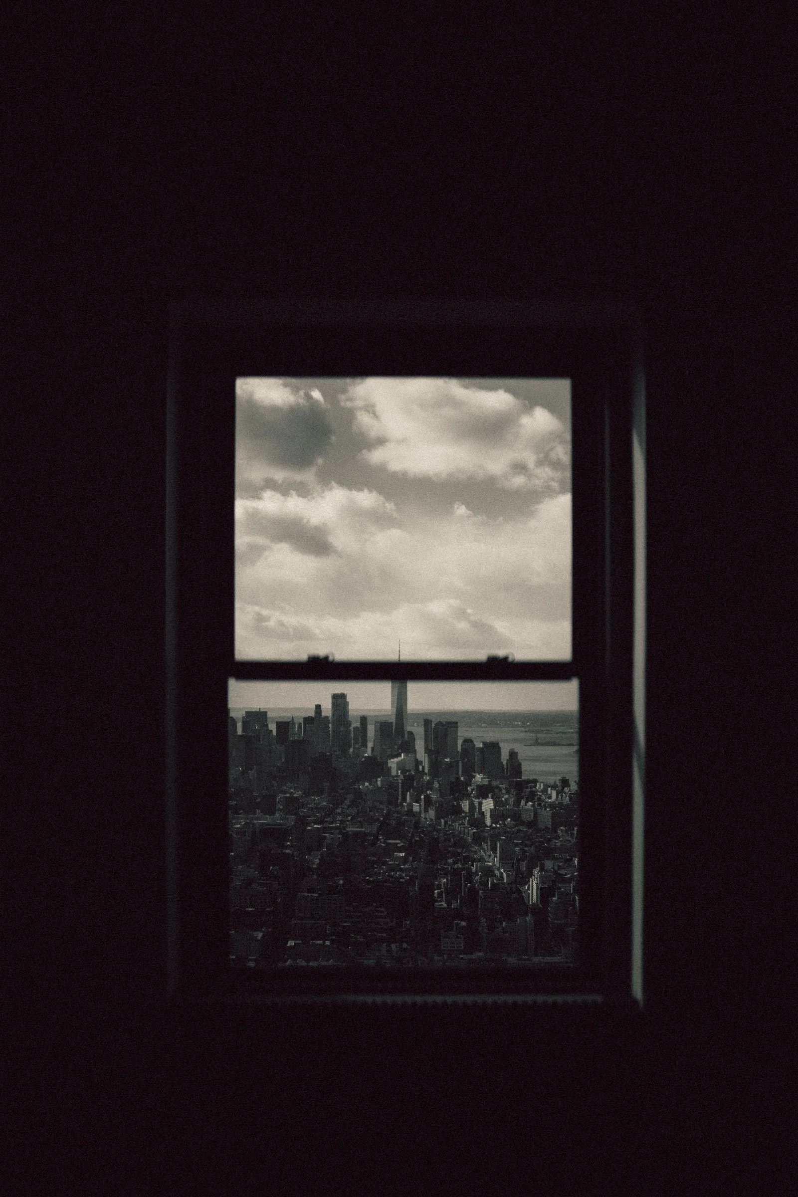 A black-and-white photo of a cityscape seen through a window with a grid, showing skyscrapers and a partly cloudy sky.