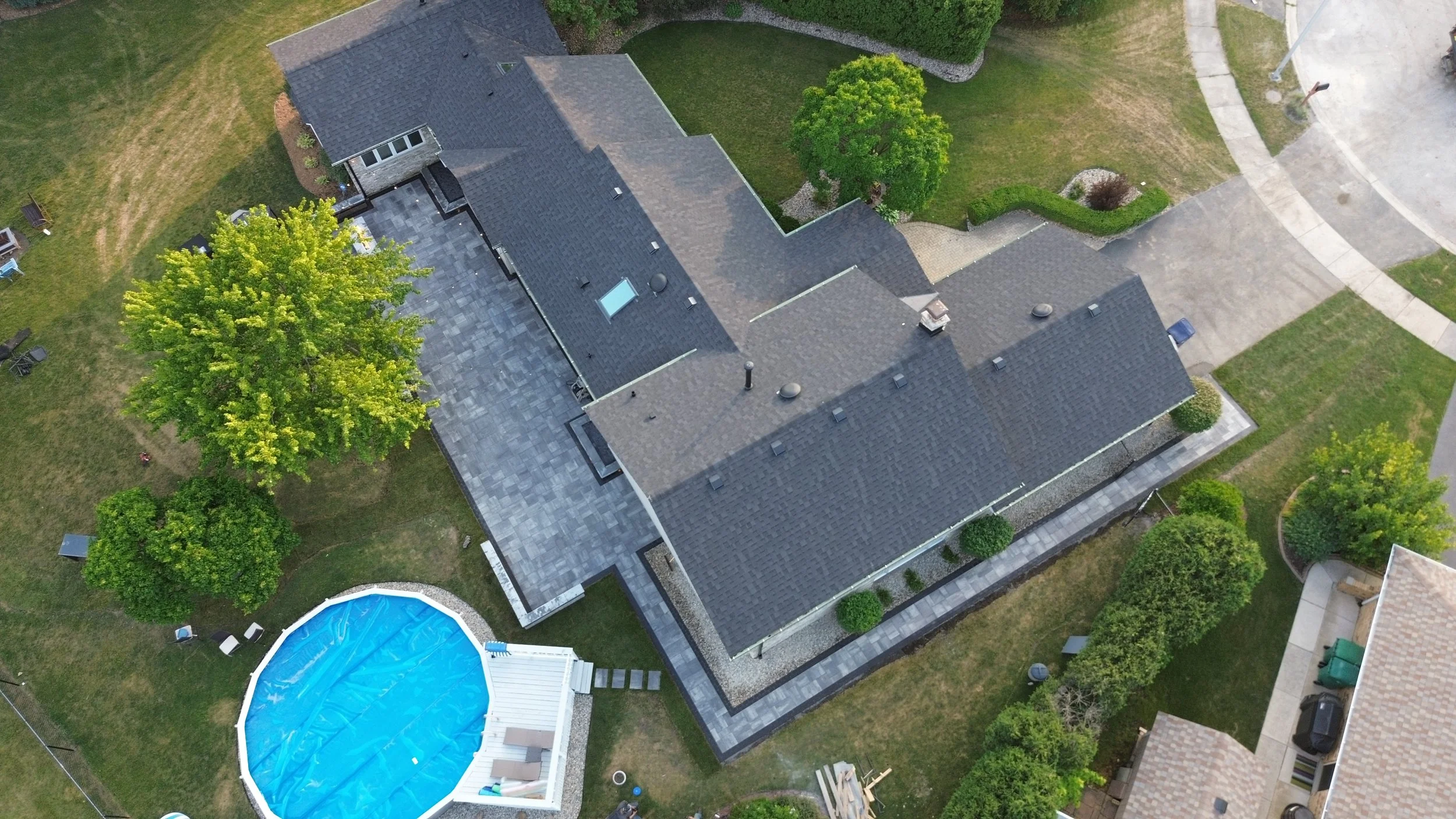 An aerial view of a residential house with a dark gray roof, a backyard with green trees, shrubs, and a round swimming pool covered with a blue tarp.