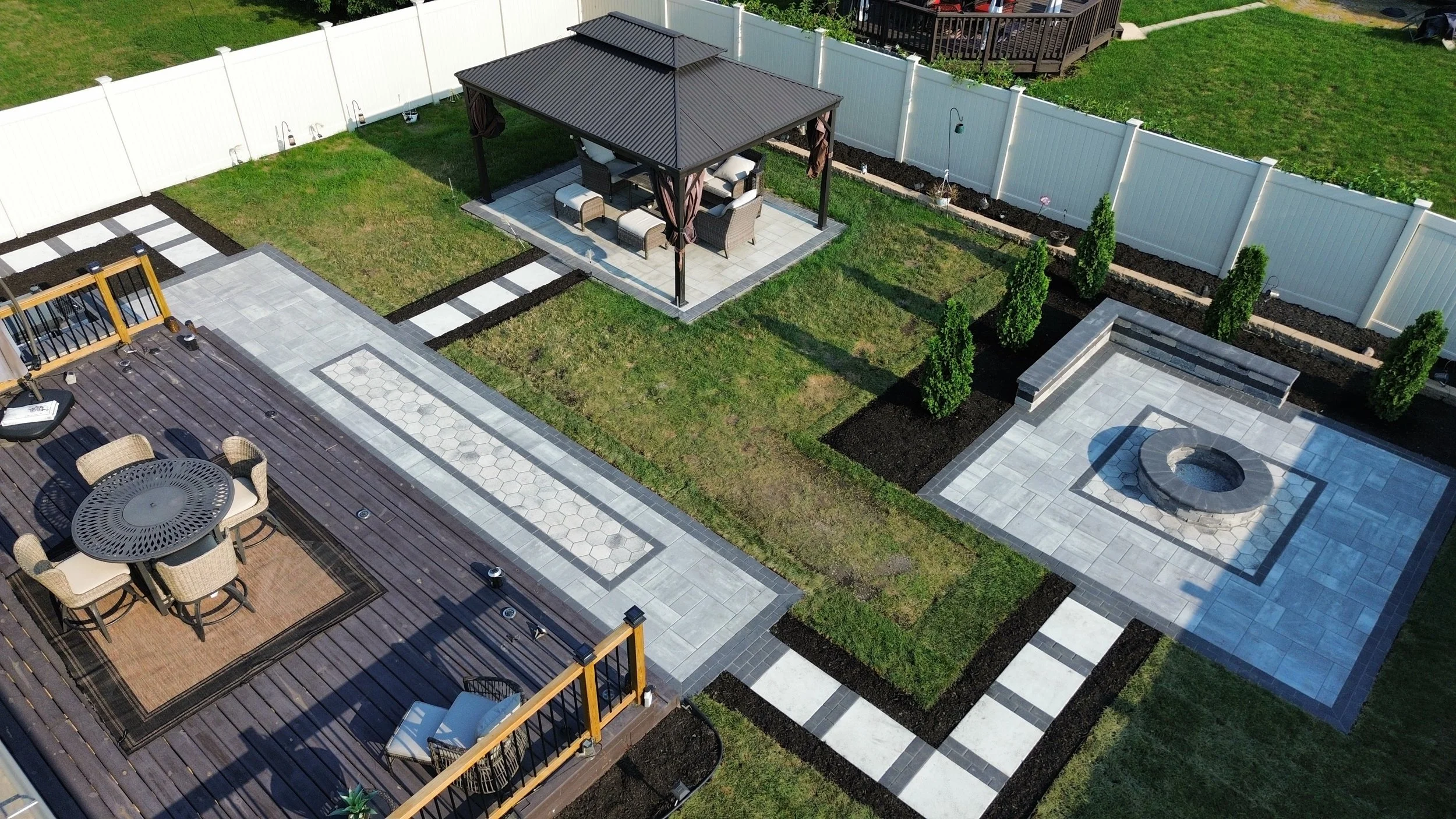 Aerial view of a backyard with a wooden deck, outdoor dining table with chairs, a sitting area with a gazebo, a fire pit, and landscaped lawns with shrubs and a white fence.