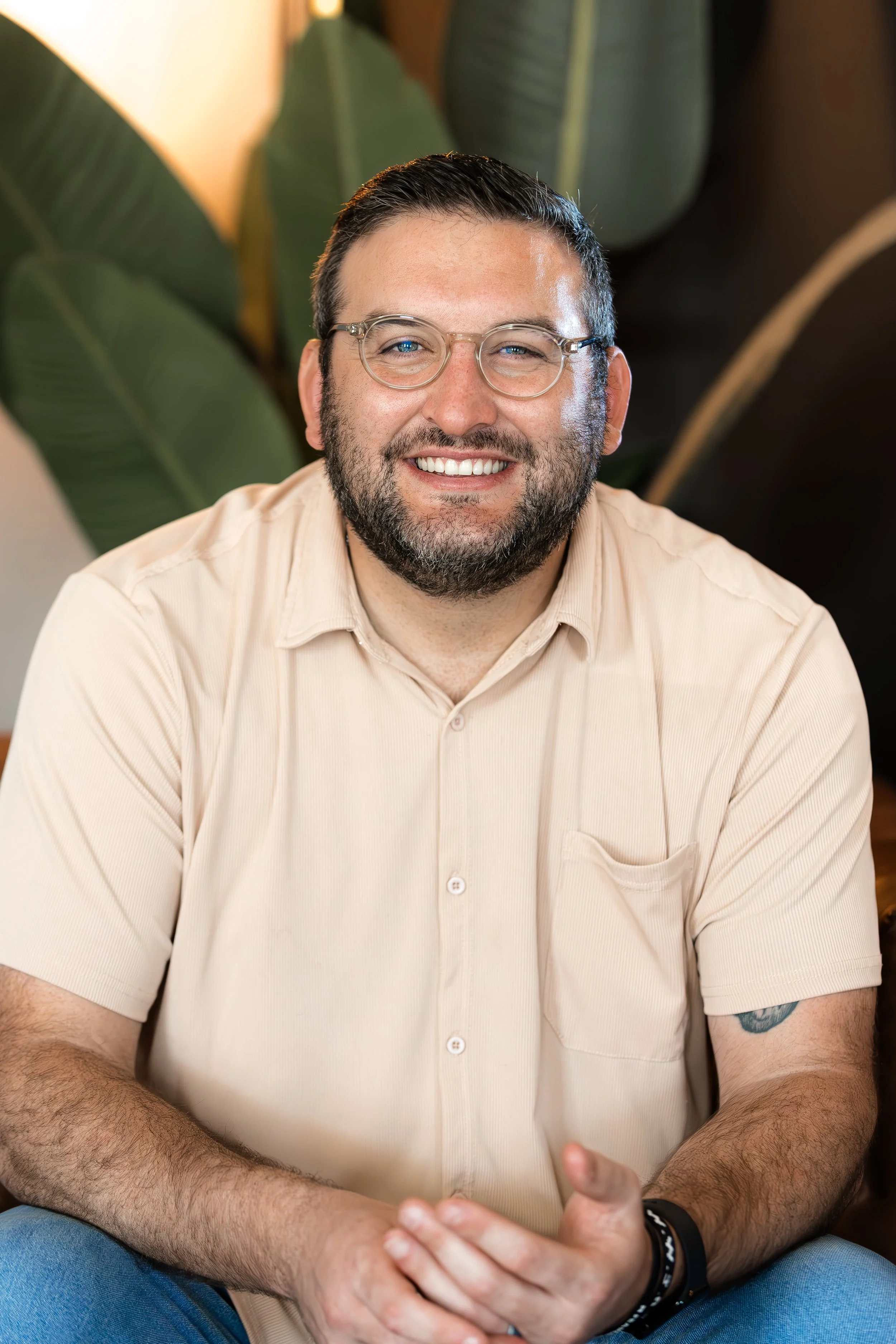 A smiling man with glasses, a beard, and short dark hair sitting indoors with large green plants in the background.