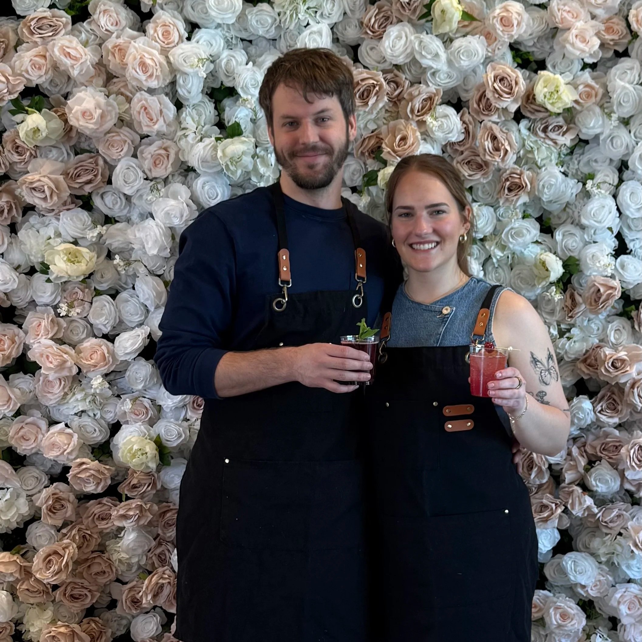 A man and woman smiling and holding drinks in front of a wall of white, cream, and pink roses.