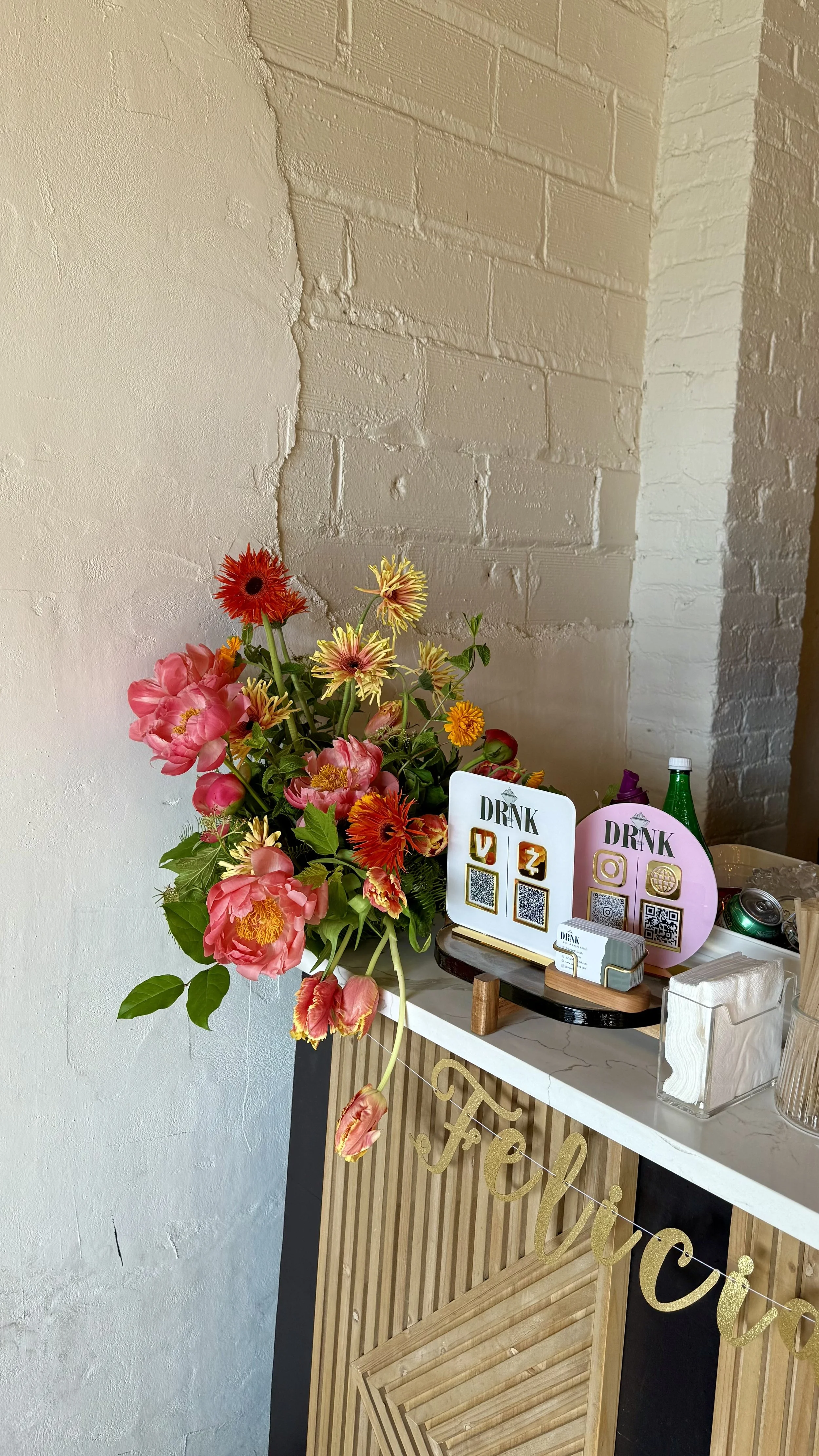 A flower arrangement with pink, orange, and yellow flowers on a table beside drink signs with QR codes, and party supplies in the background, against a brick wall with a peeling section.
