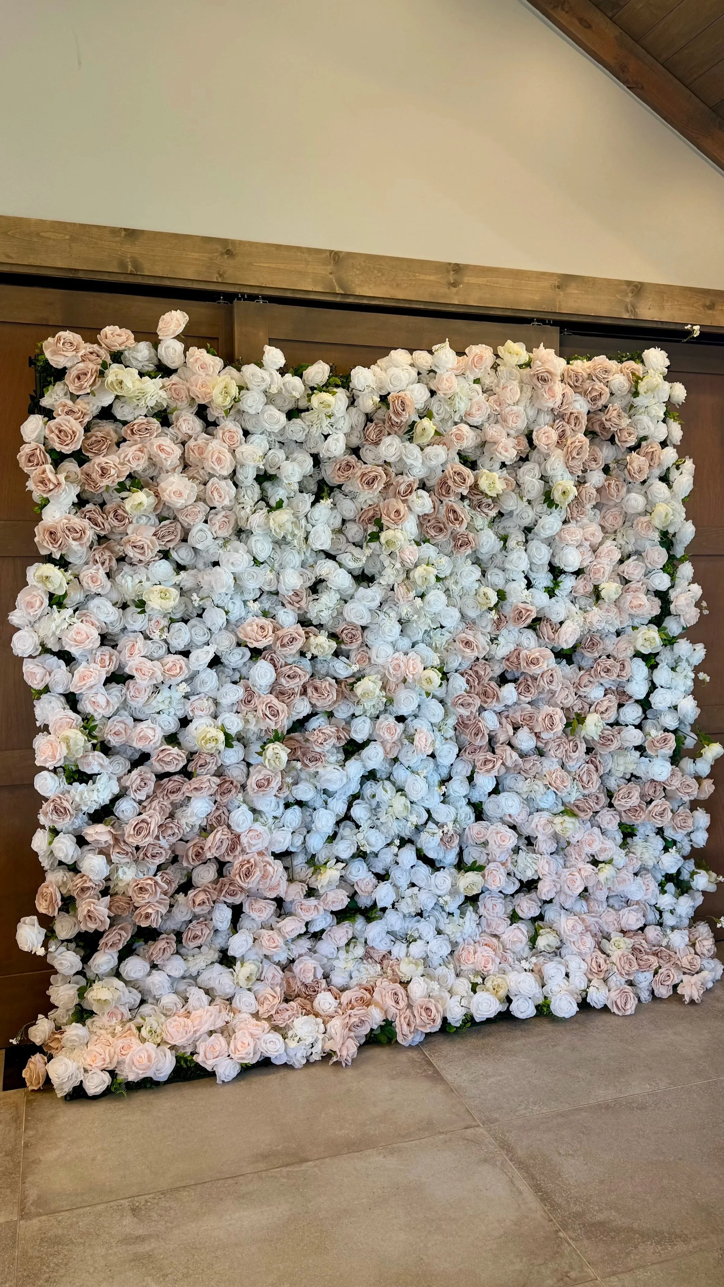 A large decorative wall covered in white, blush pink, and cream roses, set against a wooden wall in an indoor space.