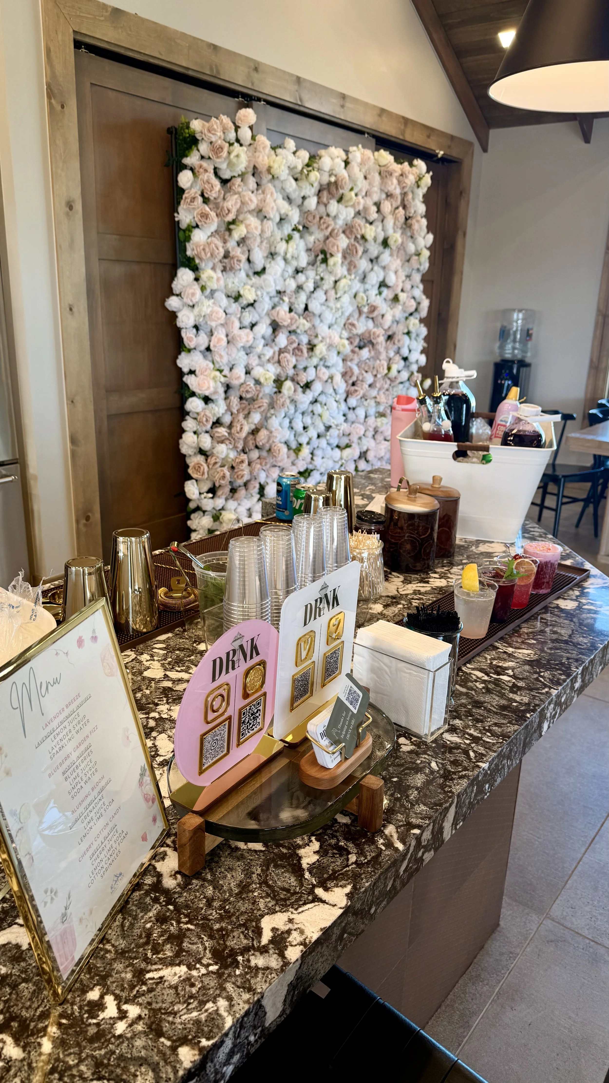 A bar area with a floral backdrop of white and light pink flowers, a marble countertop with various drinks, cups, bottles, and a drink menu, and a water dispenser in the background.