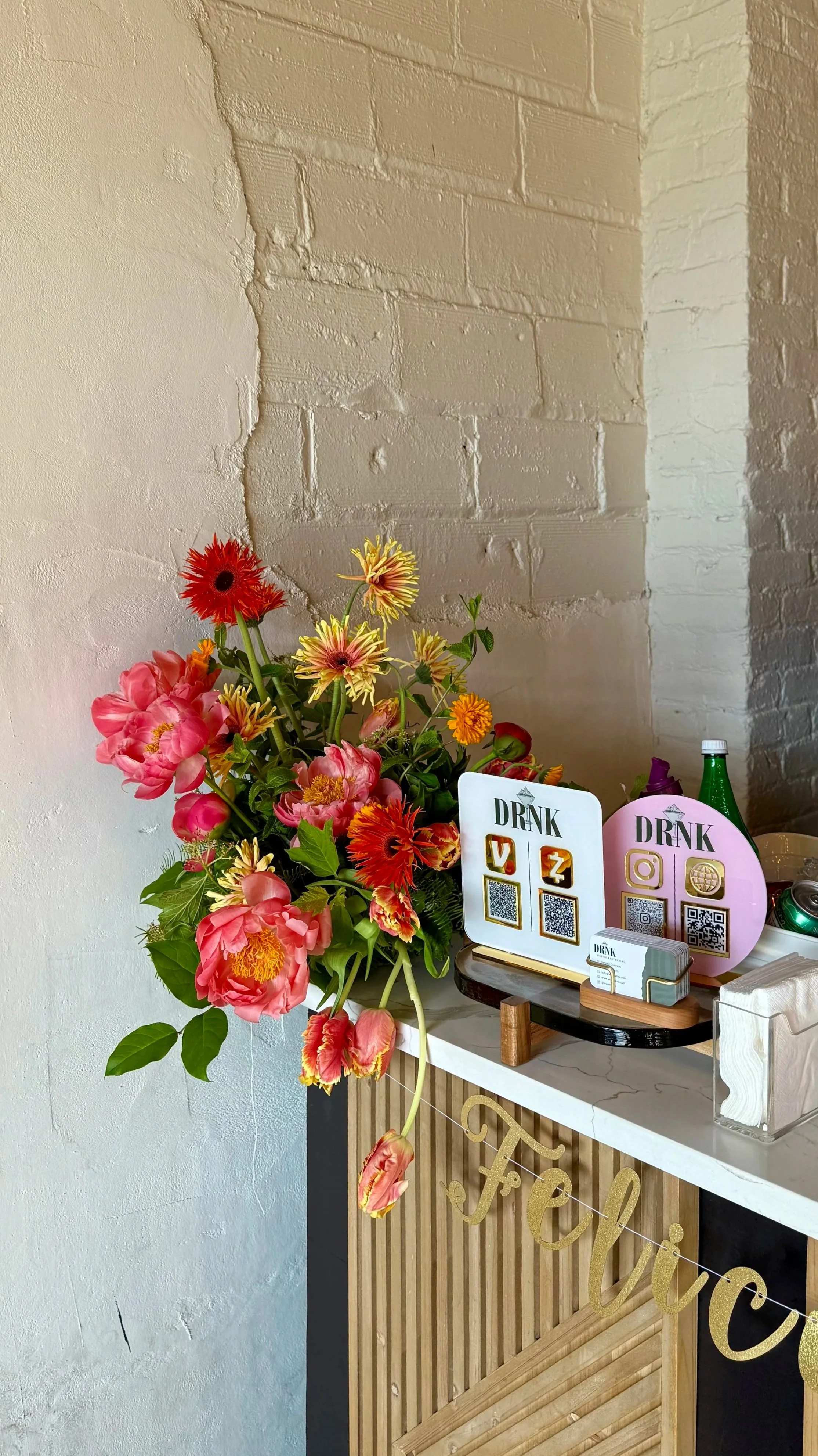 A bouquet of colorful flowers on a stand next to a wooden counter with drink QR codes, small signs, and decorations in a cafe or event setting.